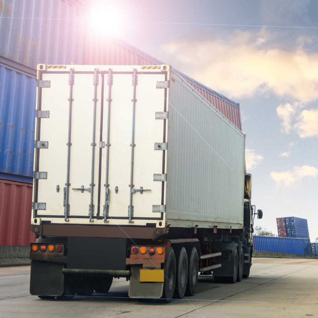 A large semi-truck with a white cargo container is parked in an industrial area among stacked shipping containers, ready for spot quotes. The sun shines brightly, casting light on the scene, while the sky remains partly cloudy.