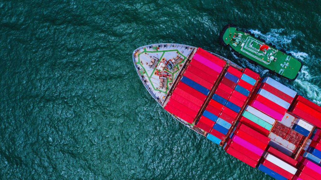 Aerial view of a large cargo ship with red and pink containers, accompanied by a small green tugboat, sailing through deep blue-green ocean water—reminding us of the documents needed for international shipping.